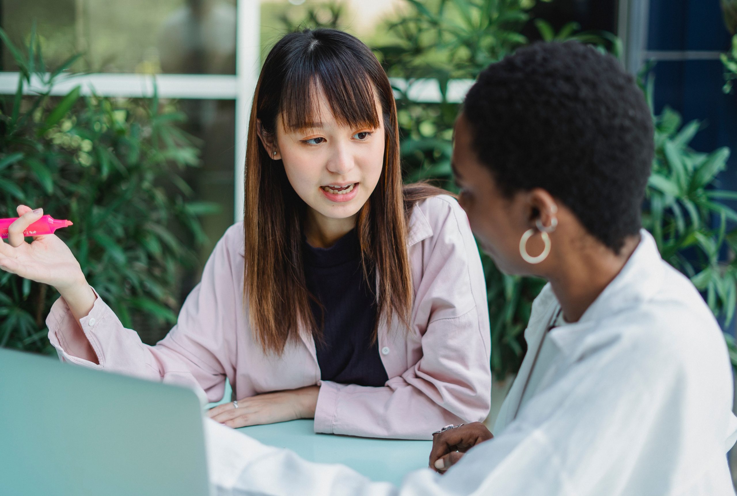 Young attentive ethnic female with marker conversing with African American partner at table with laptop while looking at each other