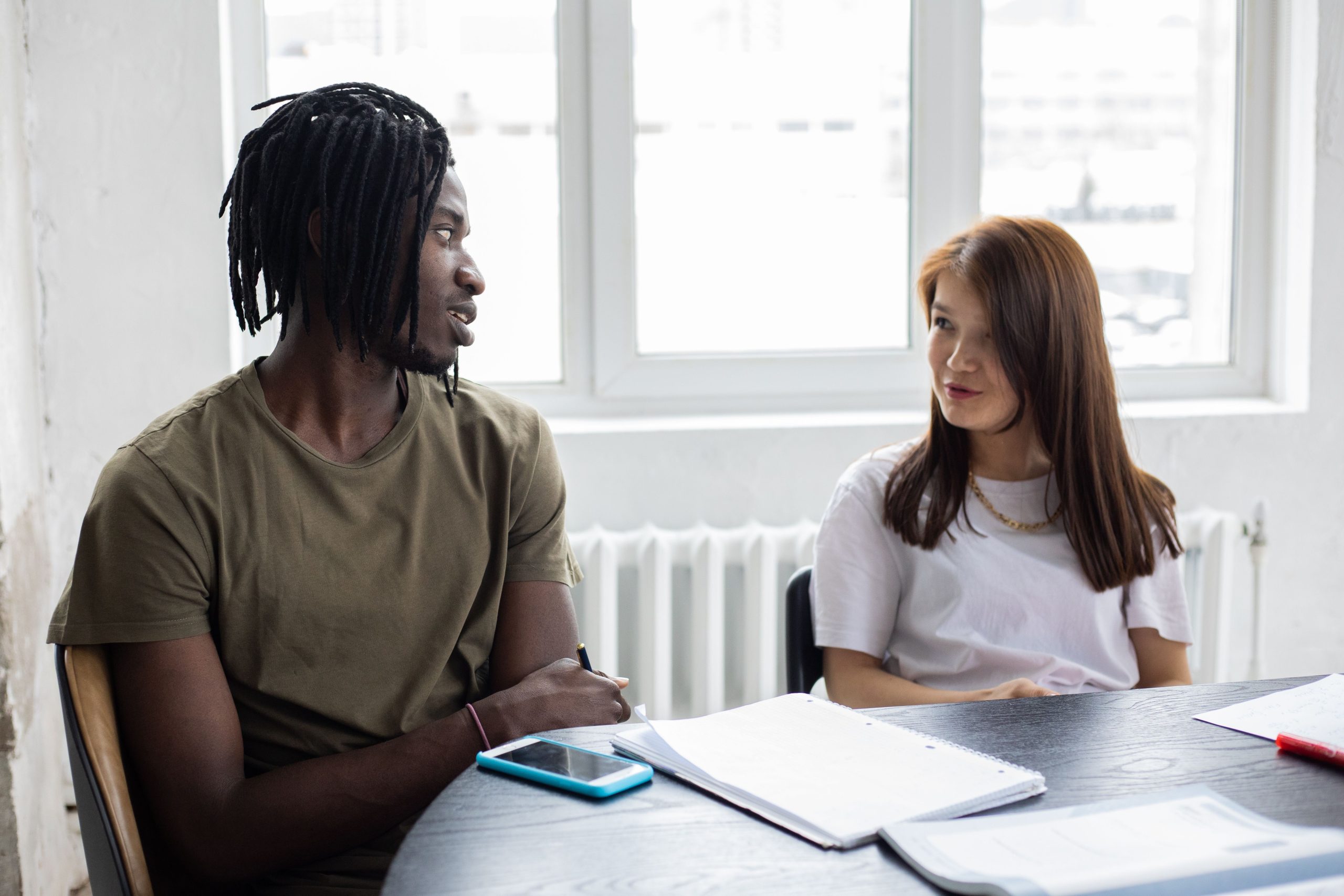 African American student and Asian female classmate chatting and looking at each other while sitting at table with notebooks during lesson