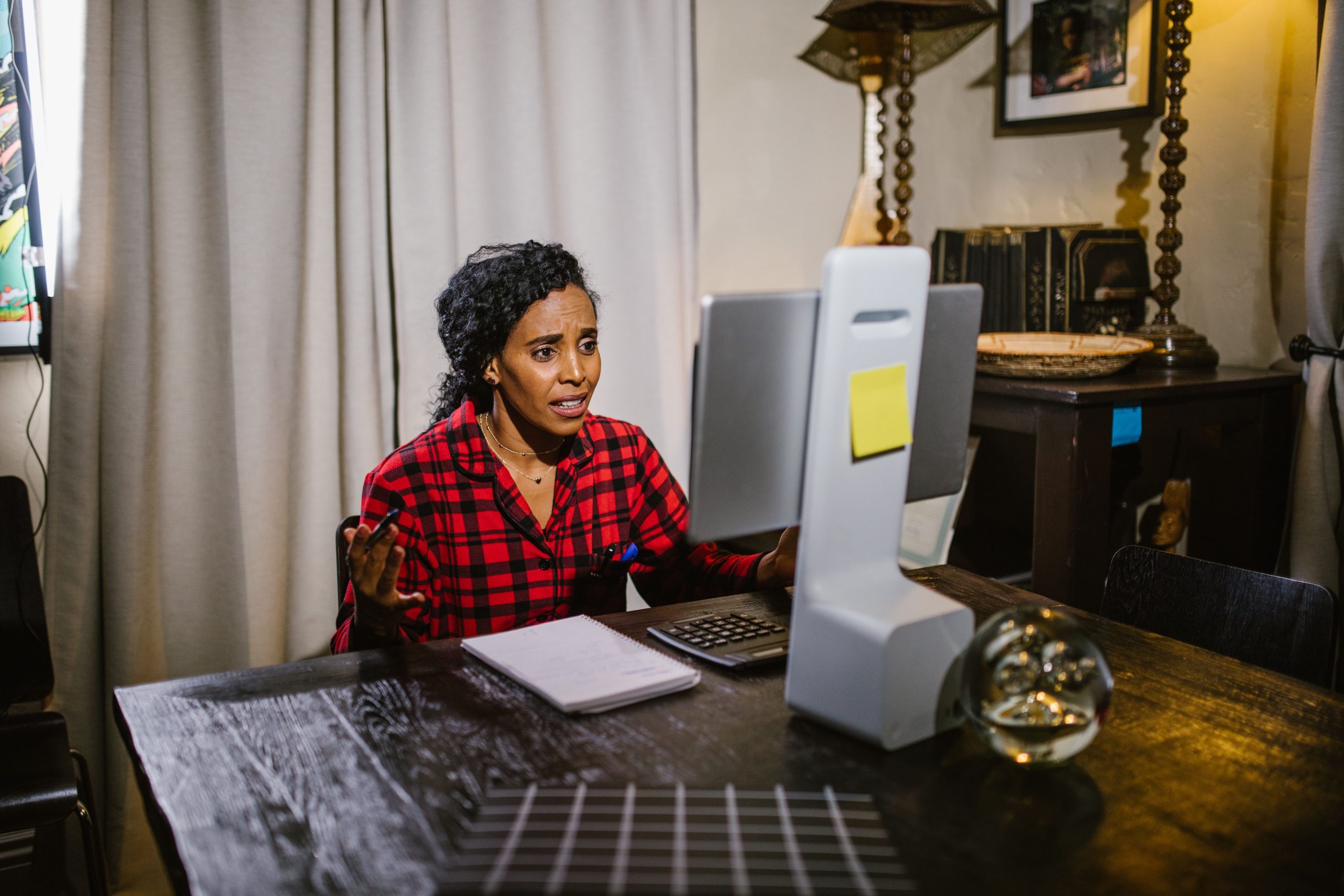 Woman in red plaid speaking at computer desk, engaged in online communication.