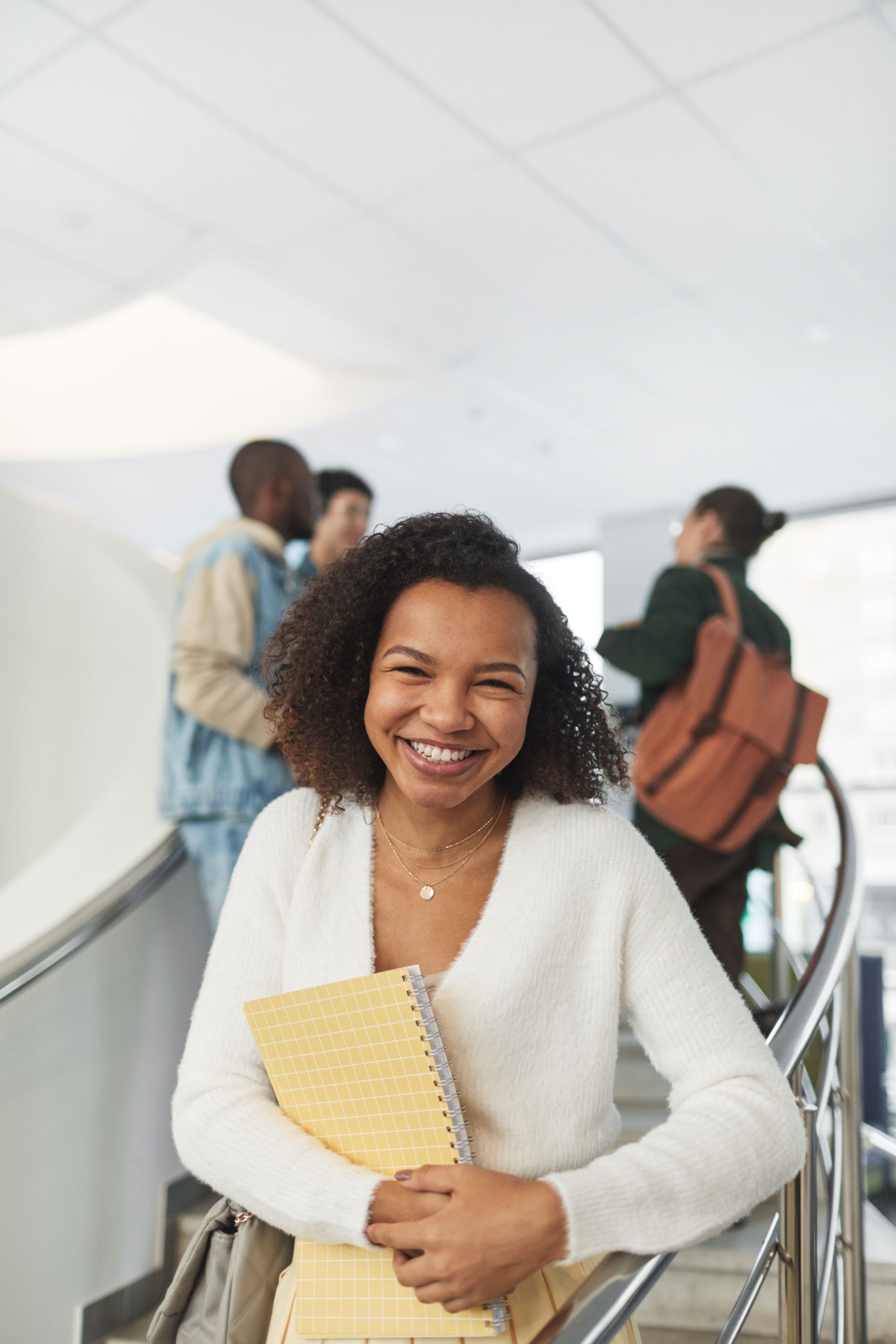 Happy student with a notebook on a staircase, surrounded by peers in an indoor educational setting.