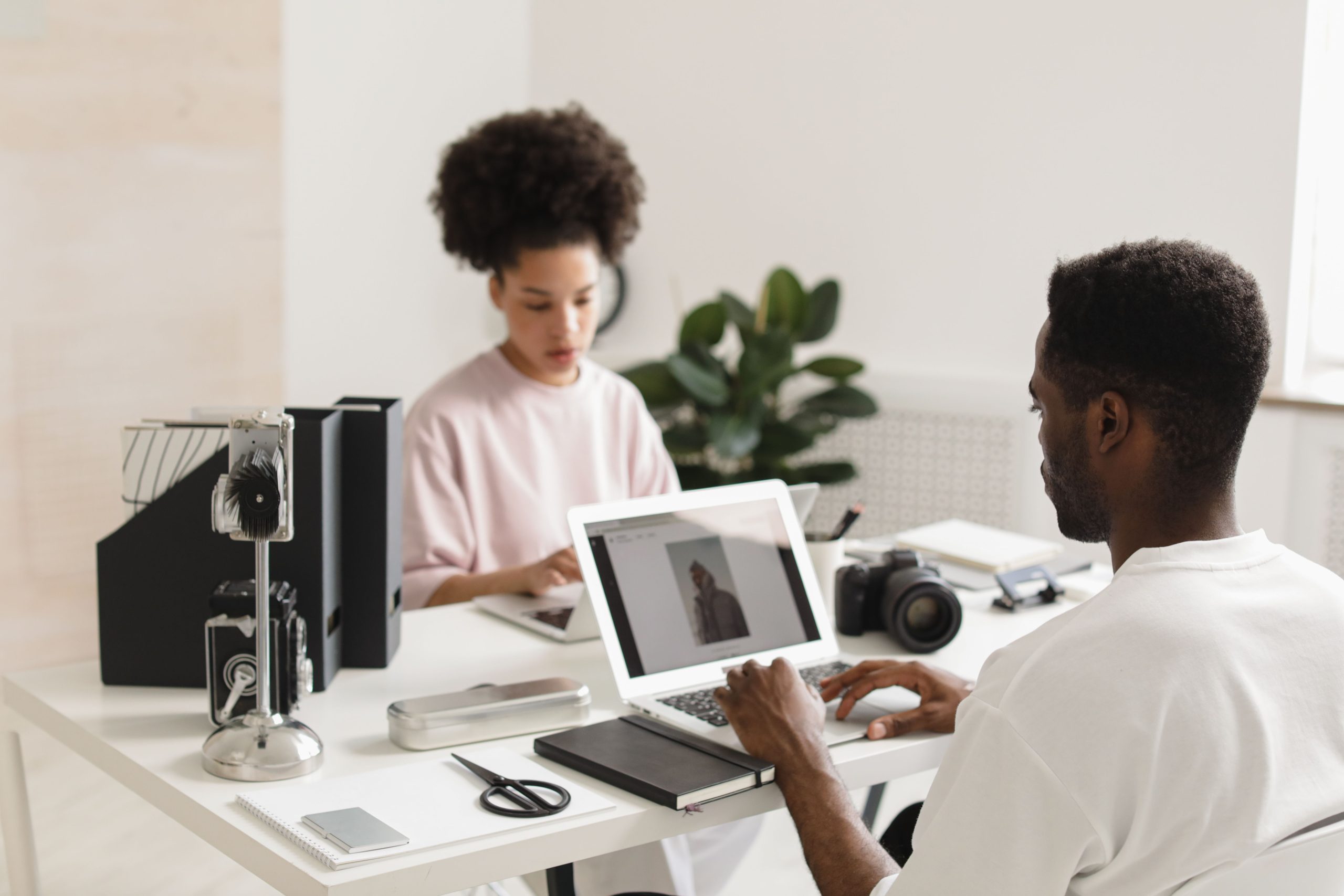 Two creative professionals collaborating in a bright, modern office setting with laptops and camera equipment.
